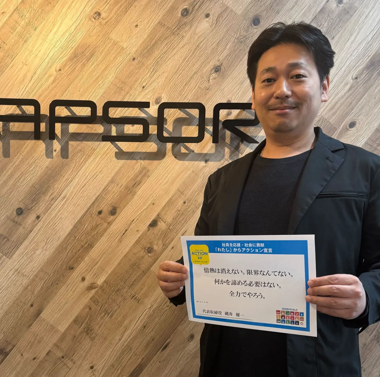 CAPSORのロゴの壁の前で、「わたし」からアクション宣言のボードを持つ代表取締役